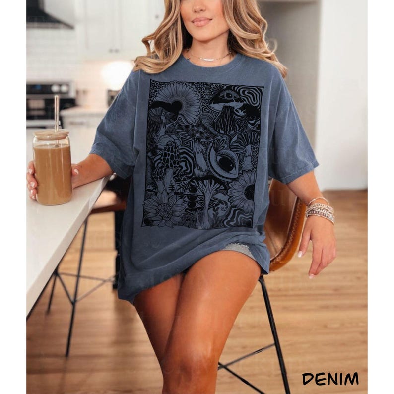 May include: A denim blue short-sleeve t-shirt featuring a large rectangular black graphic print on the front. The design consists of intricate, detailed illustrations of various mushrooms, flowers, swirling patterns, and a prominent eye.