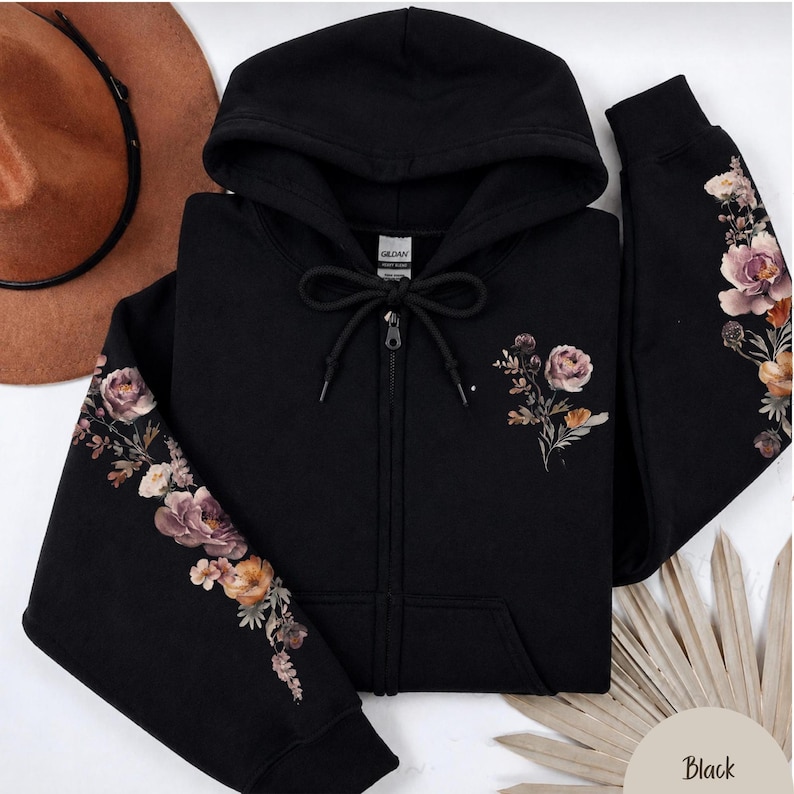 May include: A black zip-up hoodie featuring a floral print on the sleeves and the left chest. The design includes muted pink, orange, and brown flowers with green foliage. The hoodie has a drawstring hood, a full-length zipper, and ribbed cuffs.