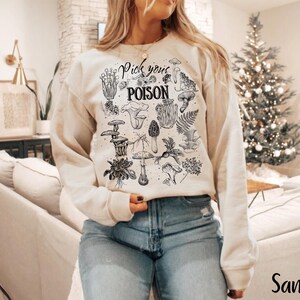 May include: Sand-colored sweatshirt featuring a black and white illustration of various mushrooms and the text "Pick your POISON". The long-sleeved top is made of a soft material and is suitable for casual wear.