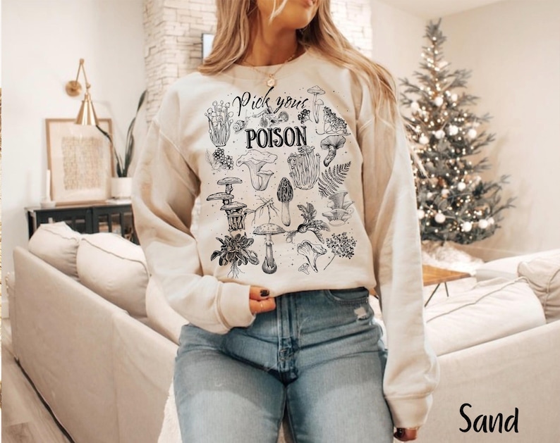 May include: Sand-colored sweatshirt featuring a black and white illustration of various mushrooms and the text "Pick your POISON". The long-sleeved top is made of a soft material and is suitable for casual wear.