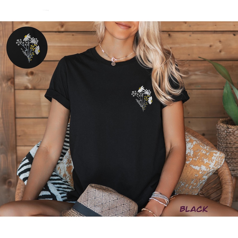 May include: Black t-shirt with a small embroidered floral design on the left chest. The design features white daisies and green stems.