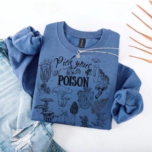 May include: A blue indigo sweatshirt with the text "Pick your POISON" and various mushroom illustrations. The sweatshirt is paired with light blue ripped jeans and a silver necklace.