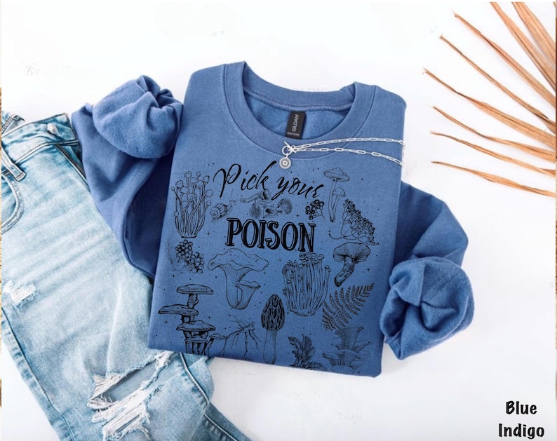 May include: A blue indigo sweatshirt with the text "Pick your POISON" and various mushroom illustrations. The sweatshirt is paired with light blue ripped jeans and a silver necklace.