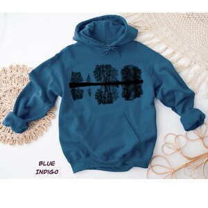 K&ouml;nnte beinhalten: Indigoblauer Kapuzenpullover mit einer schwarzen Grafik von B&auml;umen, die sich im Wasser spiegeln. Der Hoodie hat eine Kapuze mit Kordelzug und eine Vordertasche. Der Text "BLUE INDIGO" ist unten im Bild aufgedruckt.
