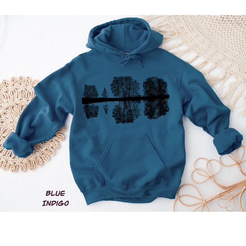 K&ouml;nnte beinhalten: Indigoblauer Kapuzenpullover mit einer schwarzen Grafik von B&auml;umen, die sich im Wasser spiegeln. Der Hoodie hat eine Kapuze mit Kordelzug und eine Vordertasche. Der Text "BLUE INDIGO" ist unten im Bild aufgedruckt.