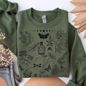 Può includere: Felpa verde militare con illustrazione botanica nera. Il design include una falena, fasi lunari e varie piante, tra cui una bottiglia, farfalle e libellule. La felpa è a girocollo.