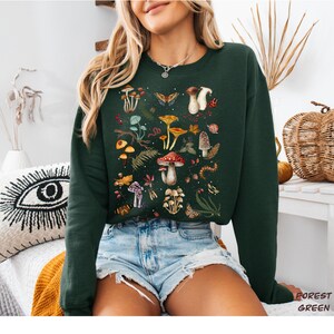 K&ouml;nnte beinhalten: Ein waldgr&uuml;ner Pullover mit einem bunten Pilz- und Naturdruck. Der Druck zeigt Pilze, Schmetterlinge, Bl&auml;tter und andere Waldelemente.