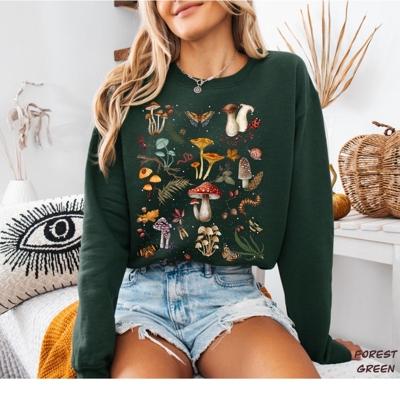 K&ouml;nnte beinhalten: Ein waldgr&uuml;ner Pullover mit einem bunten Pilz- und Naturdruck. Der Druck zeigt Pilze, Schmetterlinge, Bl&auml;tter und andere Waldelemente.