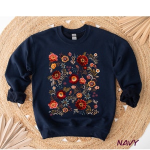 Op de afbeelding: Marineblauwe sweatshirt met een bloemenontwerp. Het geborduurde patroon heeft rode, oranje en gele bloemen met groene bladeren. Het woord "NAVY" staat rechtsonder gedrukt.