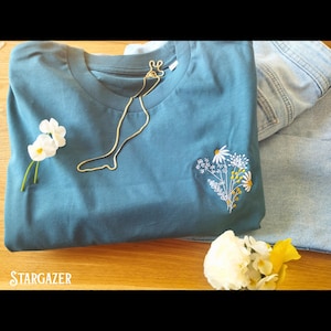May include: A teal blue t-shirt with a white embroidered floral design and a gold chain necklace. The text "STARGAZER" is visible in the bottom left corner of the image.