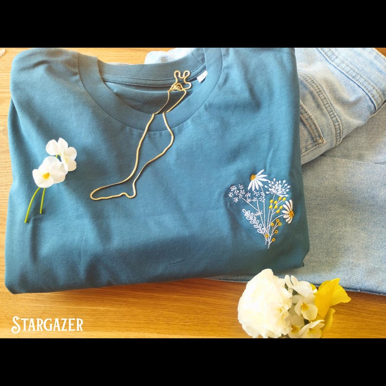 May include: A teal blue t-shirt with a white embroidered floral design and a gold chain necklace. The text "STARGAZER" is visible in the bottom left corner of the image.