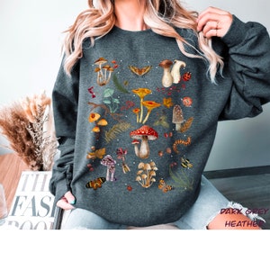 K&ouml;nnte beinhalten: Dunkelgrauer Sweatshirt mit einem Pilz- und Schmetterlings-Grafikdruck. Der Druck zeigt eine Vielzahl von Pilzen in verschiedenen Farben und Formen sowie Schmetterlinge und andere Waldelemente.