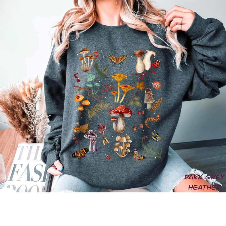 K&ouml;nnte beinhalten: Dunkelgrauer Sweatshirt mit einem Pilz- und Schmetterlings-Grafikdruck. Der Druck zeigt eine Vielzahl von Pilzen in verschiedenen Farben und Formen sowie Schmetterlinge und andere Waldelemente.