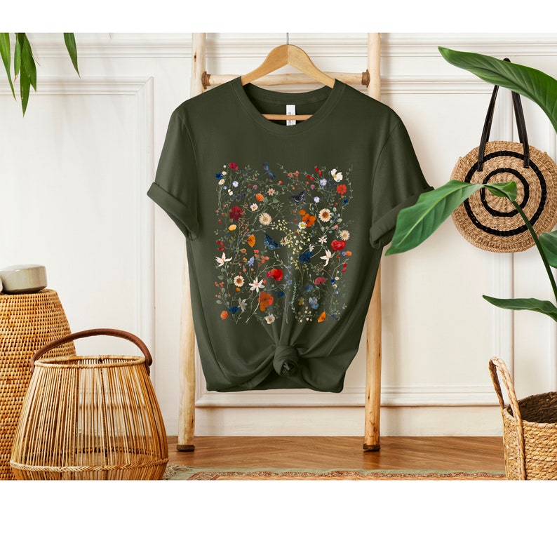 May include: A green t-shirt with a floral print featuring a variety of colorful flowers and birds. The shirt has a knotted front and is hanging on a wooden ladder.