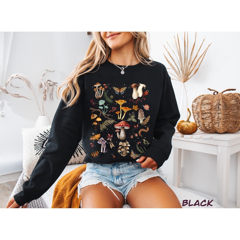 K&ouml;nnte beinhalten: Schwarzes Sweatshirt mit einem bunten Druck von Pilzen und Schmetterlingen. Der Druck zeigt verschiedene Pilzarten, Schmetterlinge und andere Waldelemente.