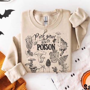 May include: A sand-colored sweatshirt with the phrase "Pick your POISON" and various mushroom illustrations in black ink. The long-sleeved top is folded, with the sleeves showing. The background includes a decorative orange blanket and black bat cutouts.