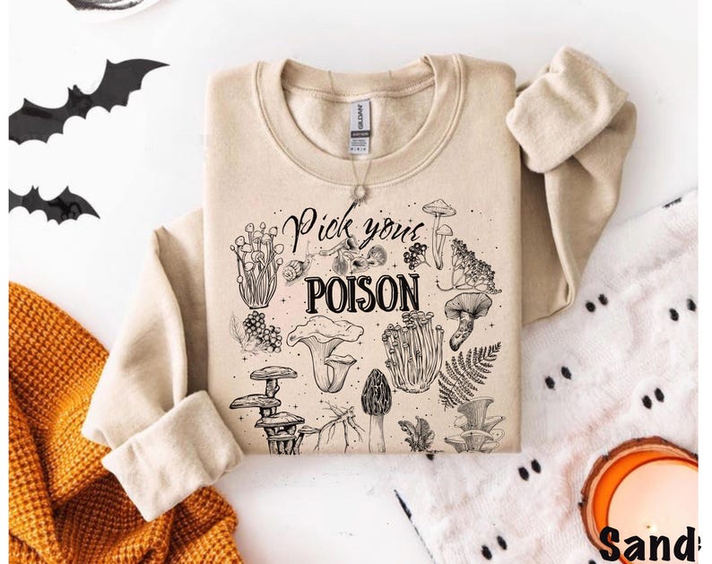 May include: A sand-colored sweatshirt with the phrase "Pick your POISON" and various mushroom illustrations in black ink. The long-sleeved top is folded, with the sleeves showing. The background includes a decorative orange blanket and black bat cutouts.