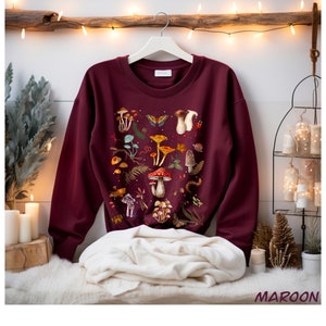 K&ouml;nnte beinhalten: Maroonfarbener Pullover mit einem bunten Druck von Pilzen und Schmetterlingen. Der Druck zeigt verschiedene Pilzarten, Schmetterlinge und andere Waldelemente.