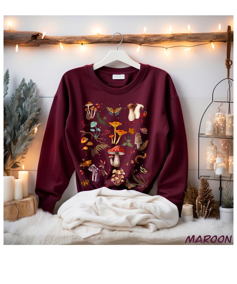 K&ouml;nnte beinhalten: Maroonfarbener Pullover mit einem bunten Druck von Pilzen und Schmetterlingen. Der Druck zeigt verschiedene Pilzarten, Schmetterlinge und andere Waldelemente.