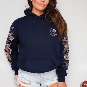 May include: A navy blue hooded sweatshirt featuring a floral print on both sleeves and a smaller floral design on the upper left chest. The hoodie includes a drawstring at the neck and a front kangaroo pocket. The fabric is a solid dark blue color.