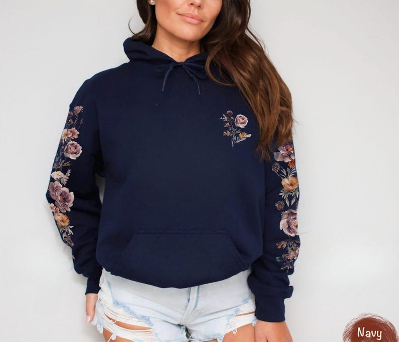 May include: A navy blue hooded sweatshirt featuring a floral print on both sleeves and a smaller floral design on the upper left chest. The hoodie includes a drawstring at the neck and a front kangaroo pocket. The fabric is a solid dark blue color.