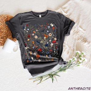 May include: A charcoal grey t-shirt with a floral print featuring a variety of flowers and birds in shades of red, orange, blue, and white.