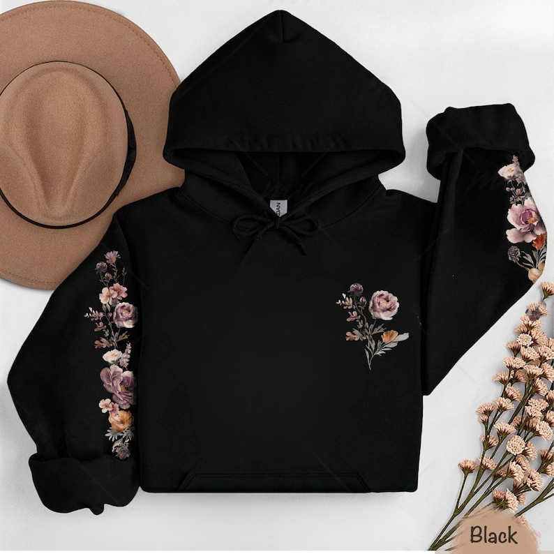 May include: A black hooded sweatshirt featuring a floral print on the left chest and along the outer length of both sleeves. The design includes muted pink, purple, and orange flowers with green foliage. The hoodie has a drawstring hood.