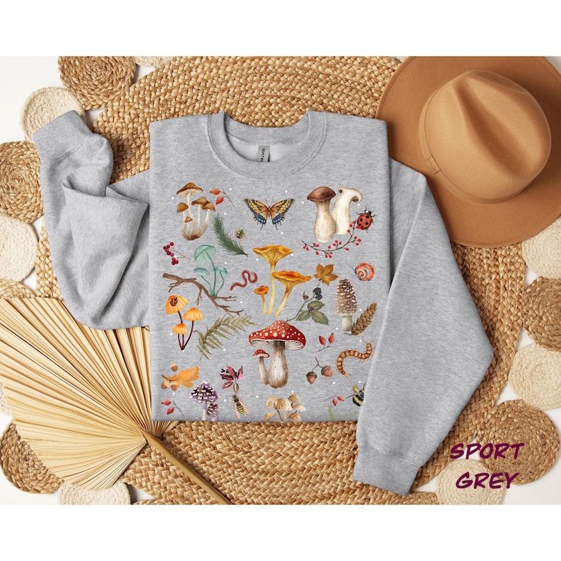 K&ouml;nnte beinhalten: Ein graues Sweatshirt mit einem bunten Pilz- und Schmetterlingsdesign. Das Design umfasst rote, orange, gelbe, braune, gr&uuml;ne und wei&szlig;e Pilze, Schmetterlinge und andere Waldelemente.