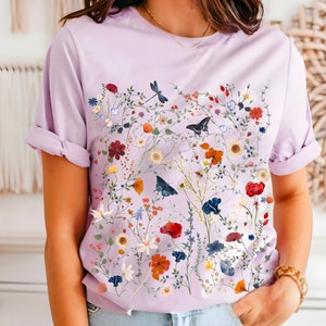 May include: A lavender t-shirt with a floral print featuring a variety of flowers, leaves, and butterflies. The print is in shades of red, orange, yellow, blue, and green.