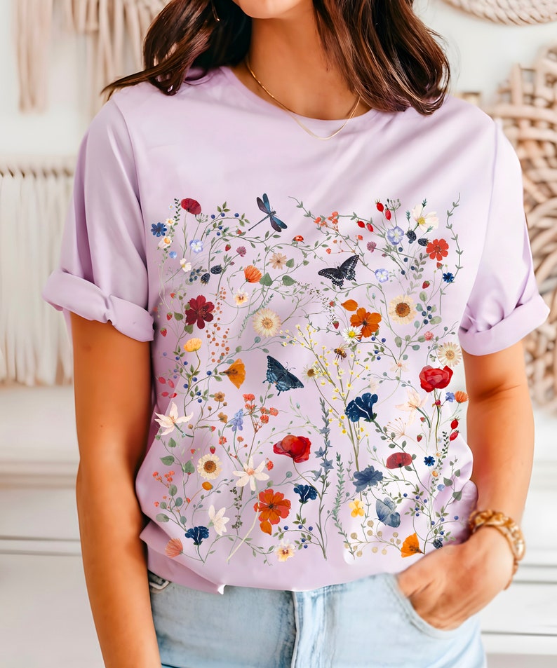 May include: A lavender t-shirt with a floral print featuring a variety of flowers, leaves, and butterflies. The print is in shades of red, orange, yellow, blue, and green.