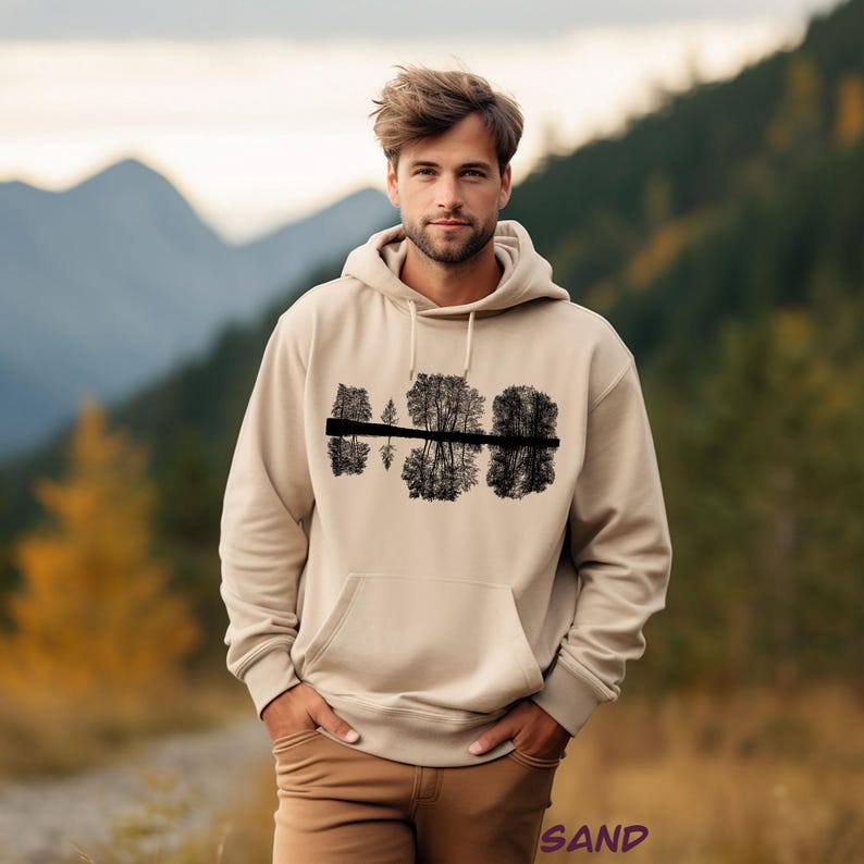 K&ouml;nnte beinhalten: Ein sandfarbener Kapuzenpullover mit einer schwarzen Grafik von B&auml;umen, die sich im Wasser spiegeln. Der Hoodie hat eine Vordertasche und eine Kapuze mit Kordelzug. Das Wort "SAND" ist unten rechts aufgedruckt.