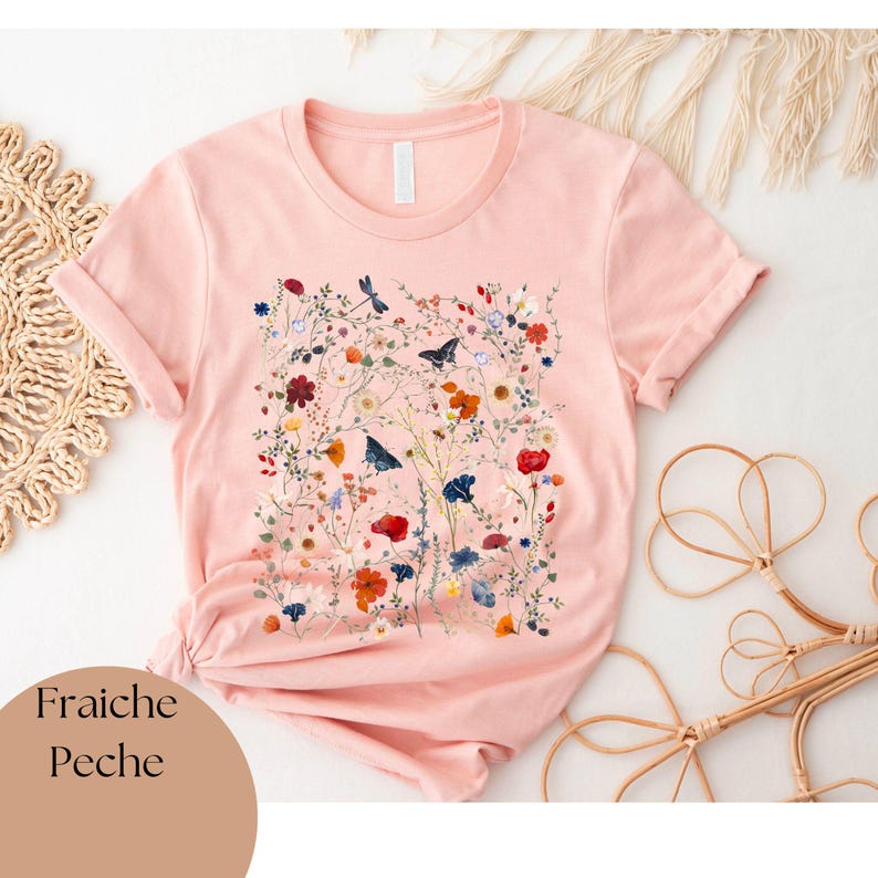 May include: A pink t-shirt with a floral print featuring a variety of flowers, leaves, and butterflies. The text "Fraiche Peche" is printed on the bottom left corner of the shirt.