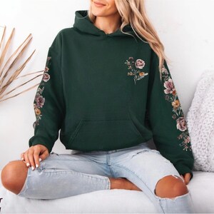 May include: A forest green hooded sweatshirt featuring a floral print on the chest and down the length of both sleeves. The design includes various flowers in shades of pink, orange, and yellow with green leaves on a dark green fabric base.
