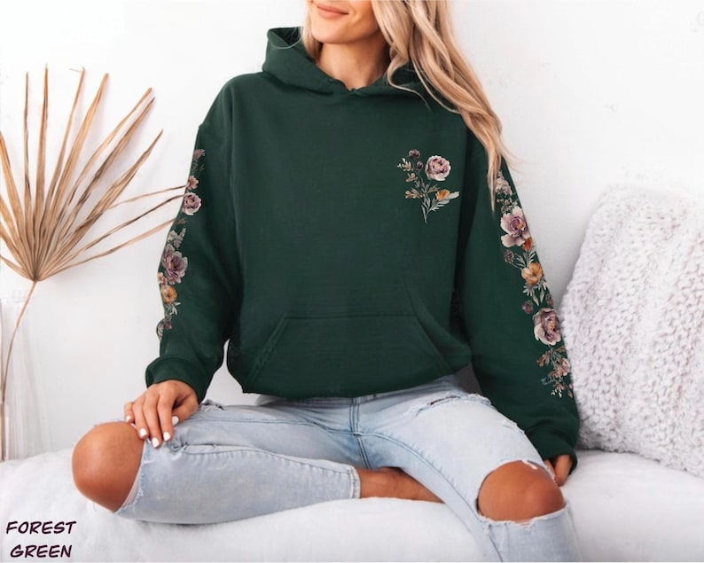 May include: A forest green hooded sweatshirt featuring a floral print on the chest and down the length of both sleeves. The design includes various flowers in shades of pink, orange, and yellow with green leaves on a dark green fabric base.