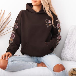 May include: A dark chocolate brown hooded sweatshirt featuring a kangaroo pocket and long sleeves. The sleeves are decorated with a floral print running down the outer length, and a smaller floral graphic is positioned on the upper left chest.