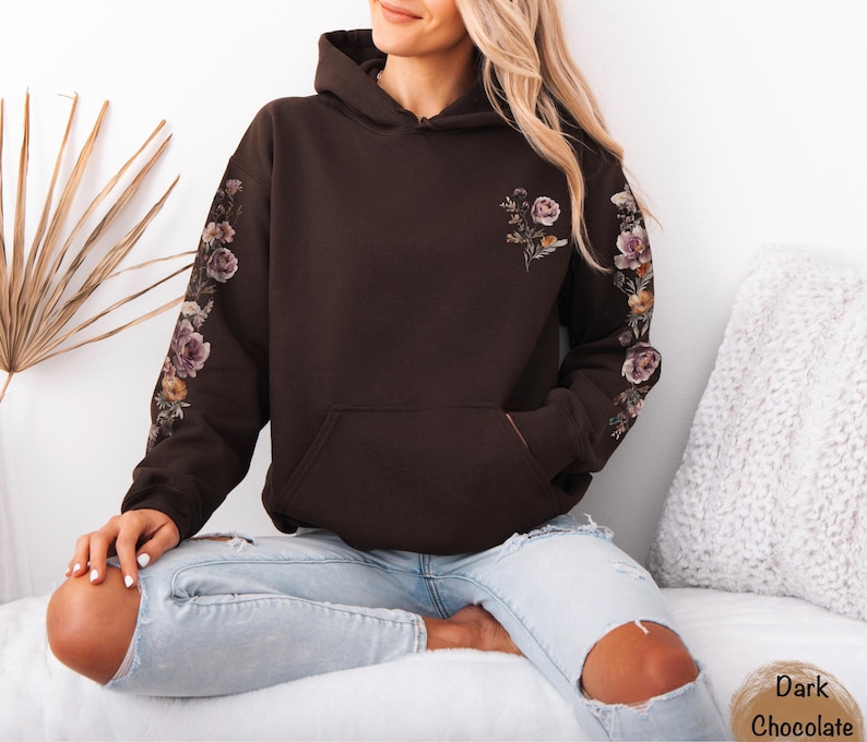 May include: A dark chocolate brown hooded sweatshirt featuring a kangaroo pocket and long sleeves. The sleeves are decorated with a floral print running down the outer length, and a smaller floral graphic is positioned on the upper left chest.