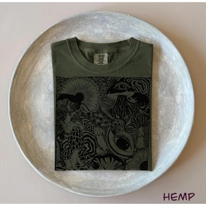 May include: An olive green t-shirt featuring a black graphic print of various mushrooms, flowers, and eyes. The shirt has a crew neckline and a tag at the inner collar that reads Comfort Colors. The design covers the center of the shirt.