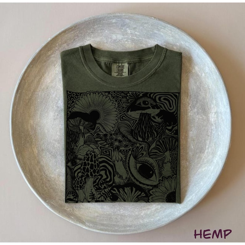 May include: An olive green t-shirt featuring a black graphic print of various mushrooms, flowers, and eyes. The shirt has a crew neckline and a tag at the inner collar that reads Comfort Colors. The design covers the center of the shirt.
