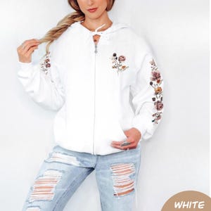 May include: A white zip-up hoodie featuring a floral print on the left chest and down the length of the sleeves. The hoodie includes a drawstring hood with white laces and a full-length front zipper closure, paired with light blue distressed jeans.