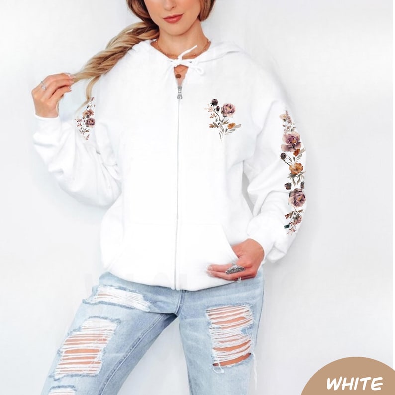 May include: A white zip-up hoodie featuring a floral print on the left chest and down the length of the sleeves. The hoodie includes a drawstring hood with white laces and a full-length front zipper closure, paired with light blue distressed jeans.