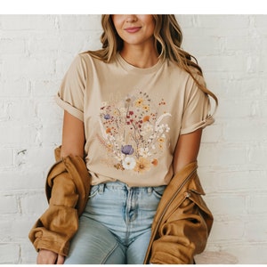 May include: A beige t-shirt with a floral print featuring a variety of flowers in shades of yellow, orange, red, purple, and white.