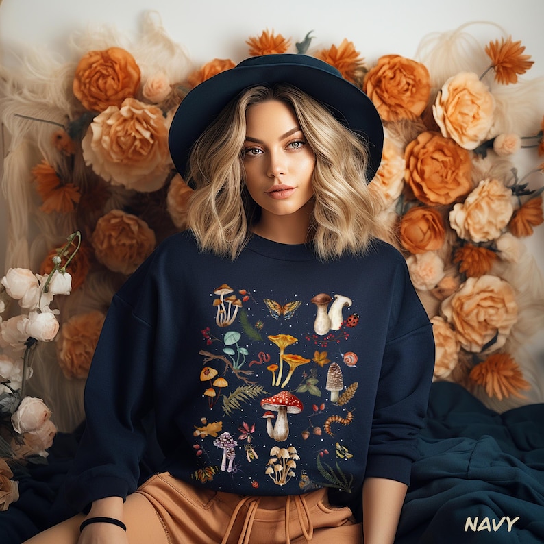 K&ouml;nnte beinhalten: Ein marineblauer Sweatshirt mit einem Pilz-Grafikdruck. Der Druck zeigt verschiedene Pilzarten, Bl&auml;tter und Insekten. Der Text "Shroom Queens" ist auf der Vorderseite des Sweatshirts gedruckt.
