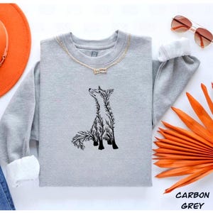 May include: A carbon grey crewneck sweatshirt featuring a black graphic of a fox composed of intricate tree branch silhouettes. The sweatshirt is displayed alongside an orange wide-brimmed hat, blue denim jeans, and brown-tinted sunglasses.