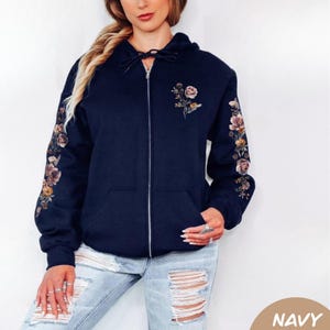 May include: A navy blue zip-up hoodie featuring a floral print on the left chest and down the length of both sleeves. The hoodie includes a drawstring hood, a full-length silver zipper, and two front pockets. The floral design consists of muted tones.