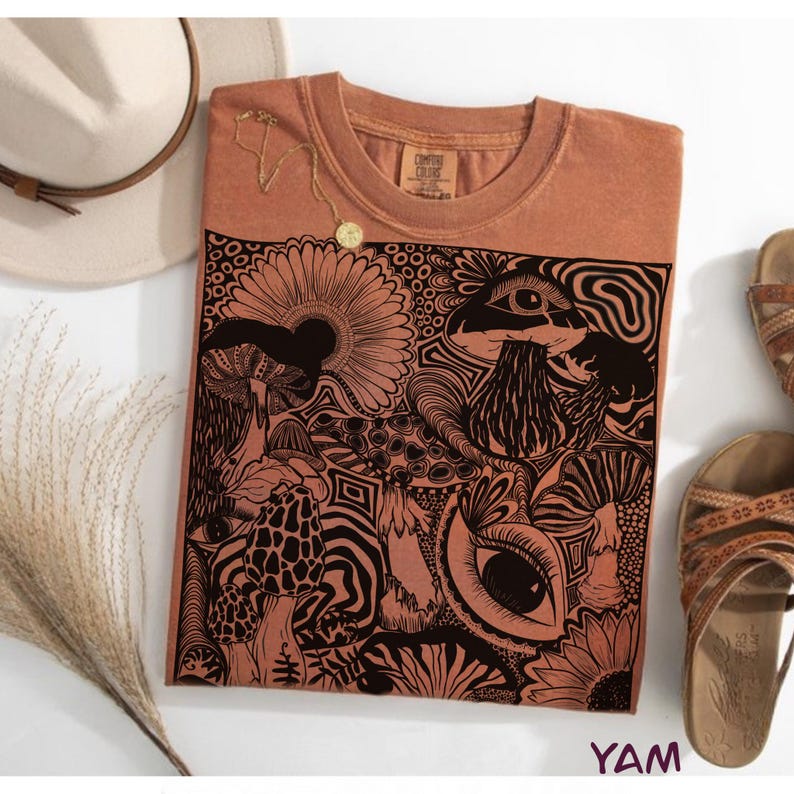 May include: A rust-colored t-shirt featuring a large black graphic print of various mushrooms, flowers, and human eyes. The shirt has a crew neckline and a tag on the inner collar. A gold-toned necklace rests on the upper chest area of the shirt.