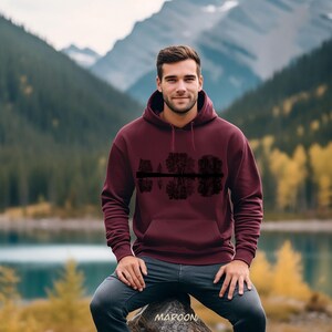 K&ouml;nnte beinhalten: Burgunderfarbener Kapuzenpullover mit einer schwarzen Grafik von B&auml;umen, die sich im Wasser spiegeln. Der Hoodie hat eine Kapuze und eine Vordertasche. Das Wort "MAROON" ist unten aufgedruckt. Der Hintergrund zeigt einen See und Berge.