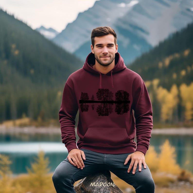 K&ouml;nnte beinhalten: Burgunderfarbener Kapuzenpullover mit einer schwarzen Grafik von B&auml;umen, die sich im Wasser spiegeln. Der Hoodie hat eine Kapuze und eine Vordertasche. Das Wort "MAROON" ist unten aufgedruckt. Der Hintergrund zeigt einen See und Berge.