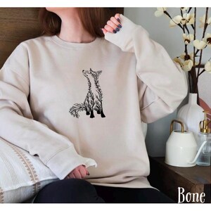 May include: A bone-colored crewneck sweatshirt featuring a black graphic illustration of a fox composed of intricate branch and leaf patterns. The long-sleeved garment has a ribbed neckline, cuffs, and hem, with the word Bone printed in the corner.