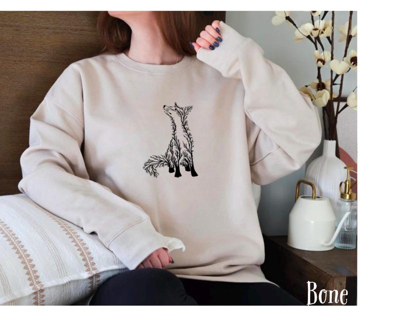 May include: A bone-colored crewneck sweatshirt featuring a black graphic illustration of a fox composed of intricate branch and leaf patterns. The long-sleeved garment has a ribbed neckline, cuffs, and hem, with the word Bone printed in the corner.