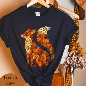 Può includere: Una t-shirt blu navy con un design di volpe realizzato con fiori pressati. La volpe è delineata da fiori arancioni, gialli e bianchi.